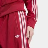 Pantalón deportivo Adidas Firebird Pinstripe color rojo con finas rayas verticales y las clásicas tres tiras blancas en los laterales. Cuenta con tiro medio, bolsillos laterales con cierre y el logo del trifolio bordado en blanco.