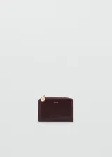 Billetera pequeña de diseño minimalista en color bordó, con cierre de cremallera y tirador metálico esférico dorado. Presenta el logo de la marca grabado en letras doradas en la parte frontal.