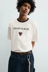 Camiseta de corte relajado confeccionada en algodón, color blanco crudo con cuello redondo en contraste color negro. Presenta un bordado frontal con la frase 'Coração de Melão' y un parche en forma de corazón.