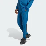 Pantalón deportivo Adidas modelo Sereno, color azul con las icónicas tres tiras en negro a los costados y logo de la marca estampado en el muslo.
