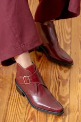Botin de cuero color bordeaux con diseño de solapa cruzada y textura grabada en el lateral. Cuenta con una hebilla metálica decorativa y taco cuadrado.