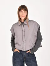 Chaleco puffer corto de color gris, con cuello camisero y cierre frontal oculto. Presenta un diseño holgado y sin mangas.