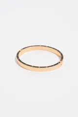 Pulsera rígida de metal dorado con detalles de esmalte negro en el borde y letras grabadas a lo largo de la pieza. Cuenta con cierre de bisagra.