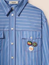 Camisa corta celeste a rayas blancas con bordes sin rematar, bolsillos de parche con solapa y pines removibles con motivos de Jamaica.