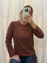 Remera de manga larga color crema, confeccionada en algodón, con cuello redondo y logo bordado en el centro del pecho. Presenta costuras decorativas en contraste en los puños y el ruedo.