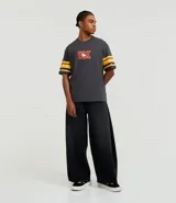 Remera gris oscuro de algodón con corte boxy, cuello redondo y mangas cortas con rayas amarillas. Estampado trasero de Hello Kitty jugando al baloncesto con letras rojas.