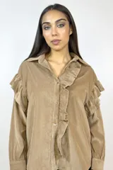 Camisa de pana fina color beige, con volados decorativos en los hombros y a lo largo de la botonadura frontal. Presenta cuello clásico, mangas largas con puños y cierre mediante botones dorados.