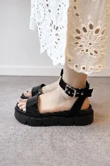 Sandalias negras de plataforma gruesa con tira ancha de charol brillante sobre los dedos y tira de gamuza ajustable con hebilla metálica en el tobillo.