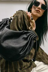 Bolso de hombro en color marrón oscuro con efecto craquelado. Presenta detalles fruncidos en los laterales con tiradores ajustables y un asa corta que combina una parte de material sintético con eslabones de cadena metálica dorada. Cuenta con un bolsillo interior y cierre mediante cremallera.