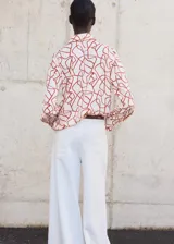 Camisa de corte recto con cuello camisero, manga larga con puños abotonados y cierre frontal de botones ocultos bajo tapeta. Presenta un diseño con estampado abstracto en tonos rojizos sobre fondo claro.