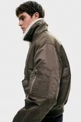 Campera bomber de nylon color marrón, ligeramente acolchada, con cuello de peluche blanco y cierre frontal. Incluye un bolsillo con cierre en una manga, bolsillos laterales con solapa y botón a presión oculto, y un bolsillo interior con botón a presión. Tiene remate acanalado ancho en puños y bajo, y un corte holgado.