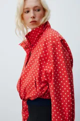 Chaqueta roja con estampado de lunares blancos, cuello subido, manga larga con puños plisados, bolsillos delanteros, bajo ajustable con cordón y cierre frontal con cremallera.