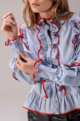 Camisa celeste con volados decorativos en los hombros, pecho, mangas y ruedo, destacados con ribetes en tono rojo. Presenta cierre frontal de botones, cuello alto y mangas largas abullonadas con puños ajustados.