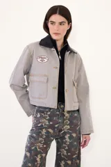 Campera corta de jean color visón, con cuello desmontable de tela engomada, cierre frontal y bolsillos plaqué con solapa en el pecho. Cuenta con un parche bordado en el delantero.