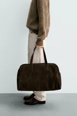 Bolso de viaje de gran tamaño confeccionado en piel con acabado de serraje. Cuenta con un compartimento principal con cierre de cremallera e imanes, interior con bolsillo con cierre y bolsillo plano, y dos asas de mano de piel.