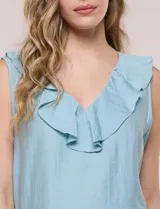 Blusa blanca sin mangas confeccionada en tejido de satén con suave textura arrugada y caída fluida. Cuenta con escote en V con volante en el cuello y ruedo estilo peplum con doble capa de volados superpuestos.