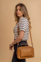 Bandolera de diseño rectangular en color suela, confeccionada en material sintético. Cuenta con un asa superior fija, correa larga ajustable y extraíble, y dos bolsillos frontales con cierre metálico, uno de ellos con diseño superpuesto. Presenta el logo de la marca grabado en el frente.