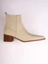 Botín de cuero sintético color beige, estilo texano, con punta fina y detalles de costuras. Tiene elásticos laterales y taco de madera.