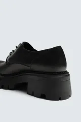 Zapato de vestir tipo derby color negro, con parte superior lisa, horma redonda y suela con plataforma track.