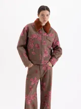 Chaqueta de gabardina color marrón con estampado floral en tonos rosados, corte recto y cierre frontal con cremallera. Presenta cuello de piel sintética en color tostado y bolsillos frontales con solapa.