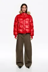 Chaqueta acolchada corta de tejido técnico brillante en color rojo, con cierre frontal de cremallera y botones de presión. Presenta capucha con borde de pelo sintético desmontable, bolsillos laterales con cremallera y puños de punto elástico en color rojo.