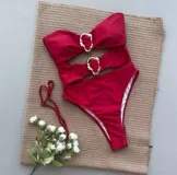 Traje de baño entero color bordó, con corte semivedetina, escote strapless y aberturas en la zona del abdomen. Presenta dos apliques metálicos dorados en forma de aro irregular.