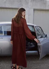 Vestido largo de tejido fluido color bordó, de diseño evasé con escote en pico y mangas largas con puños elásticos. Presenta detalles de volantes en el cuello y forro interior.