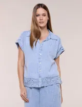 Blusa de algodón celeste con textura tramada tipo denim lavado, cuello solapa abierto en V, botones frontales y detalle de puntilla tejida al crochet en el ruedo.