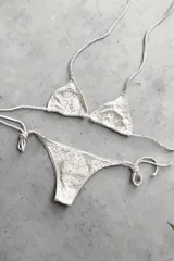 Conjunto de bikini con corpiño triangular de atar con apliques de argollitas y bombacha vedetina de atar con apliques de argollitas. El tejido presenta un estampado tie-dye o marmolado en tonos lila/azul y naranja/beige.