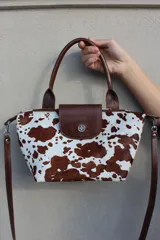 Cartera de mano con diseño de estampado vacuno, confeccionada en cuero vacuno. Cuenta con una solapa frontal con broche metálico, cierre superior, manijas cortas y correa larga desmontable.