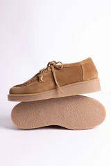 Zapato tipo mocasín acordonado de gamuza color marrón oscuro, con suela de goma color caramelo y detalle de dije metálico dorado en forma de bota colgando de los cordones.