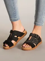 Sandalia tipo slide de color negro con diseño de tiras cruzadas y hebilla metálica ajustable. Presenta una textura grabada tipo reptil en la capellada, plantilla anatómica con borde de corcho y suela de goma plana.