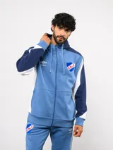 Campera deportiva Umbro del Club Nacional de Football, con cierre frontal, capucha ajustable con cordones y diseño en bloques de color azul, celeste y blanco. Presenta el logo de la marca y el escudo del club bordados en el pecho.