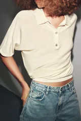 Polo de punto color crema, de manga corta y corte holgado, con cuello polo y tres botones frontales.