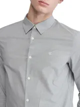 Camisa de vestir gris claro de manga larga, con cuello clásico y cierre de botones. Tiene el logo de Calvin Klein bordado en el pecho.