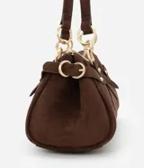 Cartera mediana de diseño alargado y estructura firme, confeccionada en gamuza sintética de color marrón. Cuenta con doble asa fina sujeta por argollas metálicas doradas, cierre superior y detalles de solapas decorativas con tachas en los laterales.