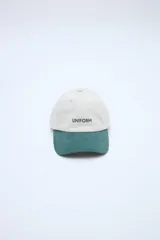 Gorra clásica con diseño en bloque de color, cuerpo en tono beige y visera en verde. Cuenta con bordado frontal con la palabra 'UNIFORM'.