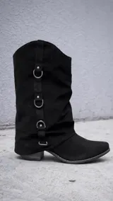 Bota de caña media estilo texano, confeccionada en gamuza negra. Presenta un diseño con efecto arrugado en la caña y una tira vertical decorativa con tres argollas metálicas plateadas. Cuenta con taco cuadrado de altura media y punta ligeramente redondeada.