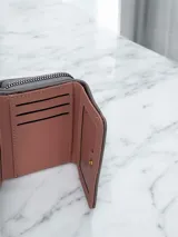 Billetera compacta de diseño plegable en color celeste, con textura granulada. Presenta una placa metálica dorada con el logo de la marca en el frente y una pequeña correa lateral del mismo tono.