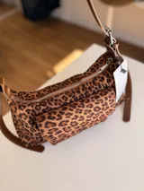 Cartera tipo baguette con estampado animal print, confeccionada en material suave símil gamuza. Presenta un bolsillo frontal con solapa y cierre, compartimento principal con cierre superior, y asa larga regulable de cuero sintético marrón.