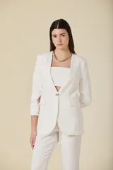 Saco de mujer color crema con diseño estructurado, cierre frontal de un botón dorado y bolsillos laterales con solapa. Presenta mangas tres cuartos con detalle de botones dorados en los puños.