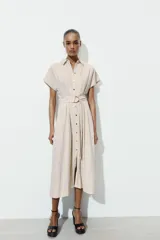 Vestido midi camisero de color beige, con cuello solapa, manga corta y cierre frontal con botones. Incluye un cinturón del mismo tejido con hebilla metálica.