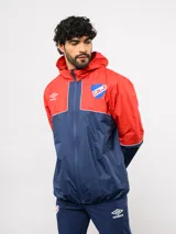 Campera de lluvia Umbro del Club Nacional de Football, con diseño de bloques de color en rojo y azul marino. Presenta capucha, cierre frontal, puños elásticos y detalles de piping blanco. Incluye logo de Umbro y escudo del club en el pecho.