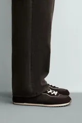 Zapatilla deportiva estilo barefoot, color marrón, con diseño de paneles en combinación de materiales y detalles en serraje. Cuenta con puntera redondeada y ancha, cordones en color crema y suela fina de goma flexible en contraste.