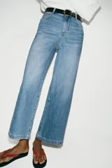 Jeans de tiro alto con pernera ancha y corte cropped. Presenta diseño de cinco bolsillos, cierre frontal con cremallera y botón, e incluye un cinturón oscuro con hebilla metálica.