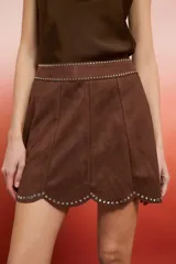 Falda corta marrón de gamuza sintética, con corte acampanado y paneles verticales. Presenta detalles de tachas plateadas a lo largo del borde superior de la cintura y en el ruedo festoneado.