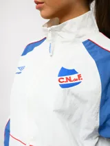 Campera deportiva Umbro del Club Nacional de Football, con diseño de bloques de color en azul marino y blanco. Presenta cuello alto, cierre frontal completo, bolsillos laterales y el escudo del club junto al logo de la marca en el pecho.