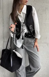 Chaleco de gamuza negra con bordados decorativos en color blanco y cierre frontal mediante cordones con borlas. Presenta un diseño corto y estilo boho.