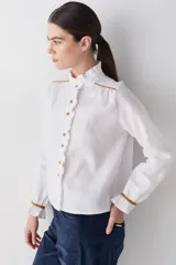 Camisa blanca de lino con cuello alto tipo gorguera, cierre frontal con botones de madera y detalles de ribetes en color mostaza en hombros y puños. Presenta volados en la botonera, cuello y puños.