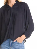 Blusa de gasa con plisado soleil, cuello a la base fruncido con escote en V regulable mediante lazos, y mangas largas abullonadas con puños sastreros.